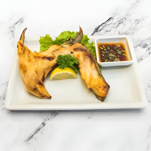 Grilled salmon collar with lemon, parsley, lettuce, and a side of dipping sauce on a white plate.